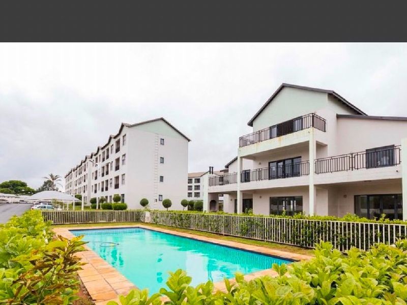 Communal swimming pool area featuring a paved surround and metal fencing, set against a multi-story residential complex with balconies.