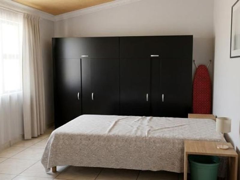 A functional bedroom featuring large built-in wardrobes, neutral tiled flooring, and natural light from a window with curtains.