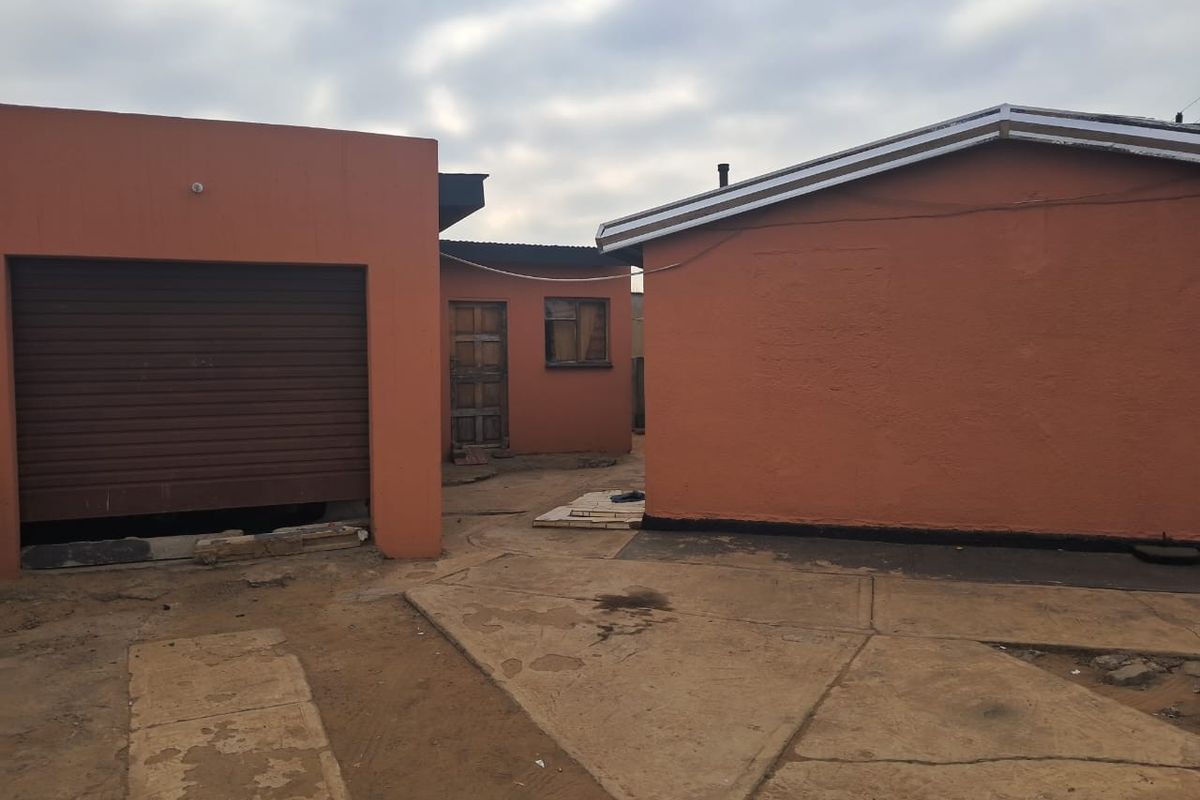 Rear exterior view of the property featuring a garage with a roller door, concrete paved courtyard, and basic building structures.