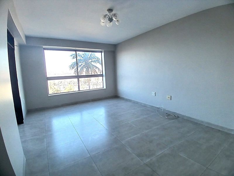 A bright, empty bedroom featuring neutral tiled flooring, a large window providing natural light, and a modern ceiling light fixture.