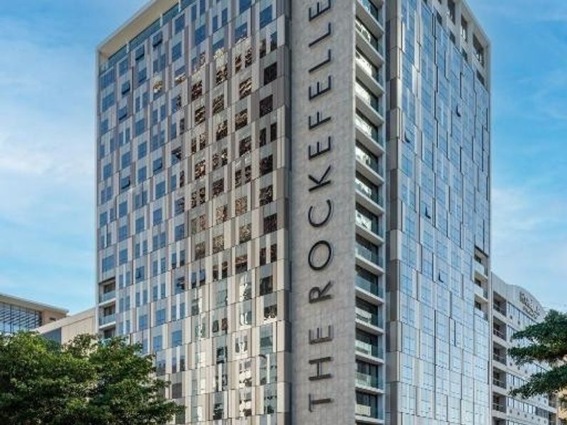 A modern high-rise apartment building featuring a glass facade, integrated balconies, and prominent building signage, situated in a busy urban environment.