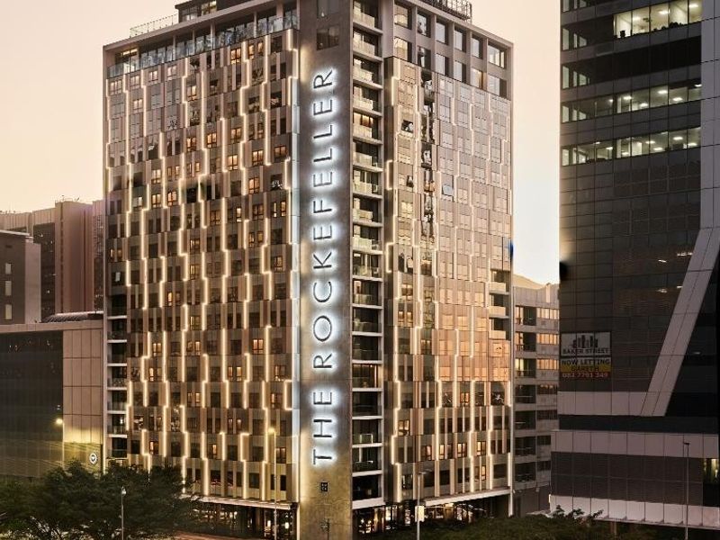 A professional exterior view of The Rockefeller apartment building, showcasing its modern architectural design, facade lighting, and urban street-level presence.