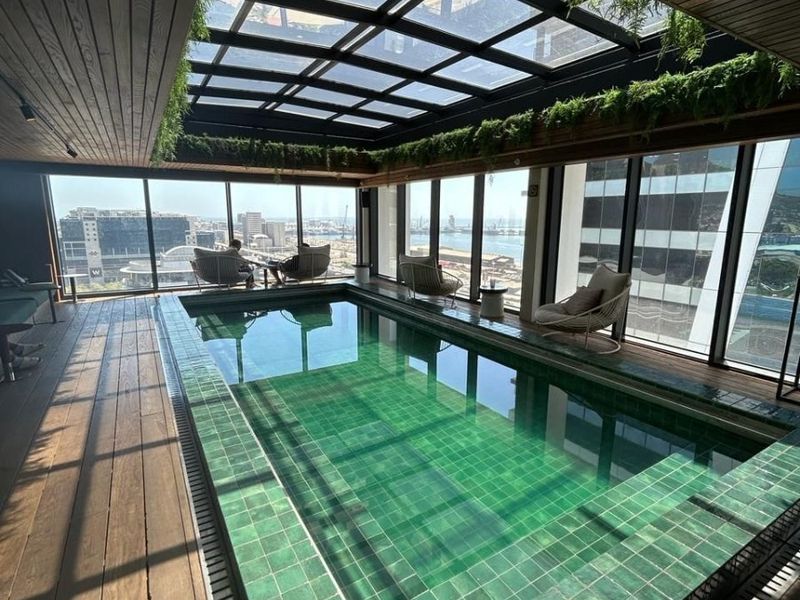A luxurious indoor swimming pool area featuring a glass skylight, timber ceiling, and floor-to-ceiling windows offering expansive city and harbour views.