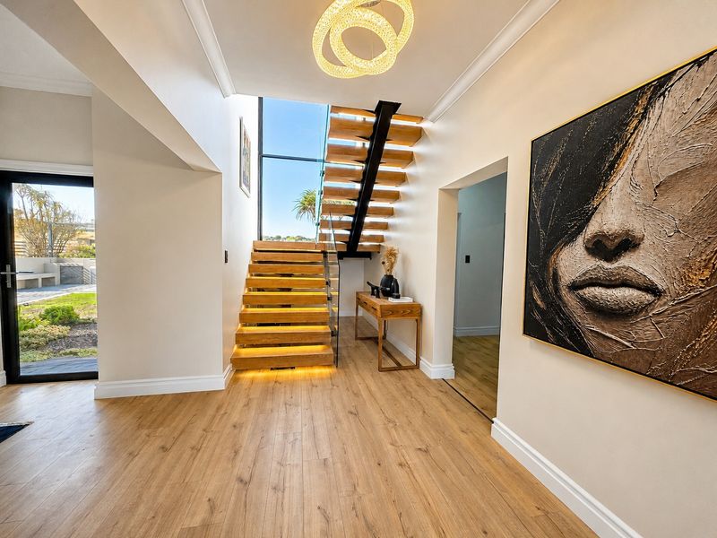 Modern entrance hall featuring a striking timber staircase with glass balustrade, integrated stair lighting, and large windows providing natural light and garden views.