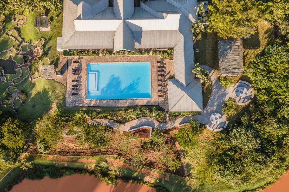 Aerial view of a property featuring a large swimming pool, expansive paved patio, landscaped gardens, and a metal roof structure.