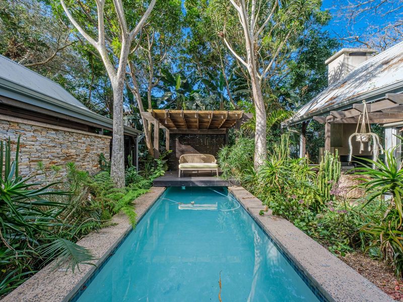 A tranquil outdoor pool area featuring a lap pool, timber decking with a pergola, stone wall accents, and lush garden surroundings.