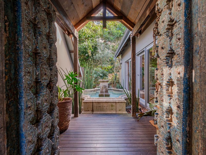 A charming covered patio featuring timber decking, a central stone water fountain, and ornate carved wooden entrance doors leading to the garden.