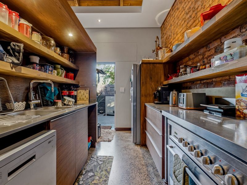 Modern industrial-style kitchen featuring exposed brick walls, stainless steel countertops, built-in oven, and polished concrete flooring with direct access to an outdoor area.