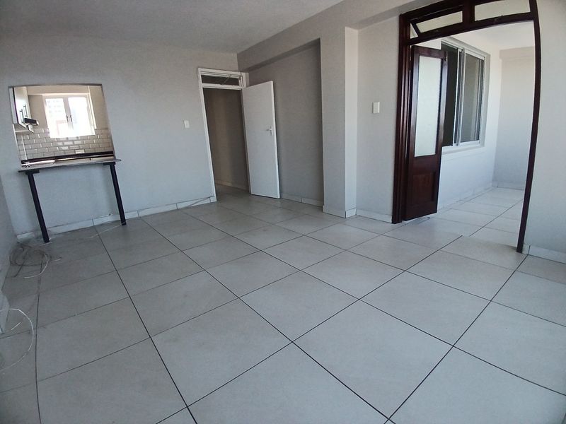 An empty open-plan living area featuring neutral tiled flooring, a kitchen pass-through window, and access to adjacent rooms.