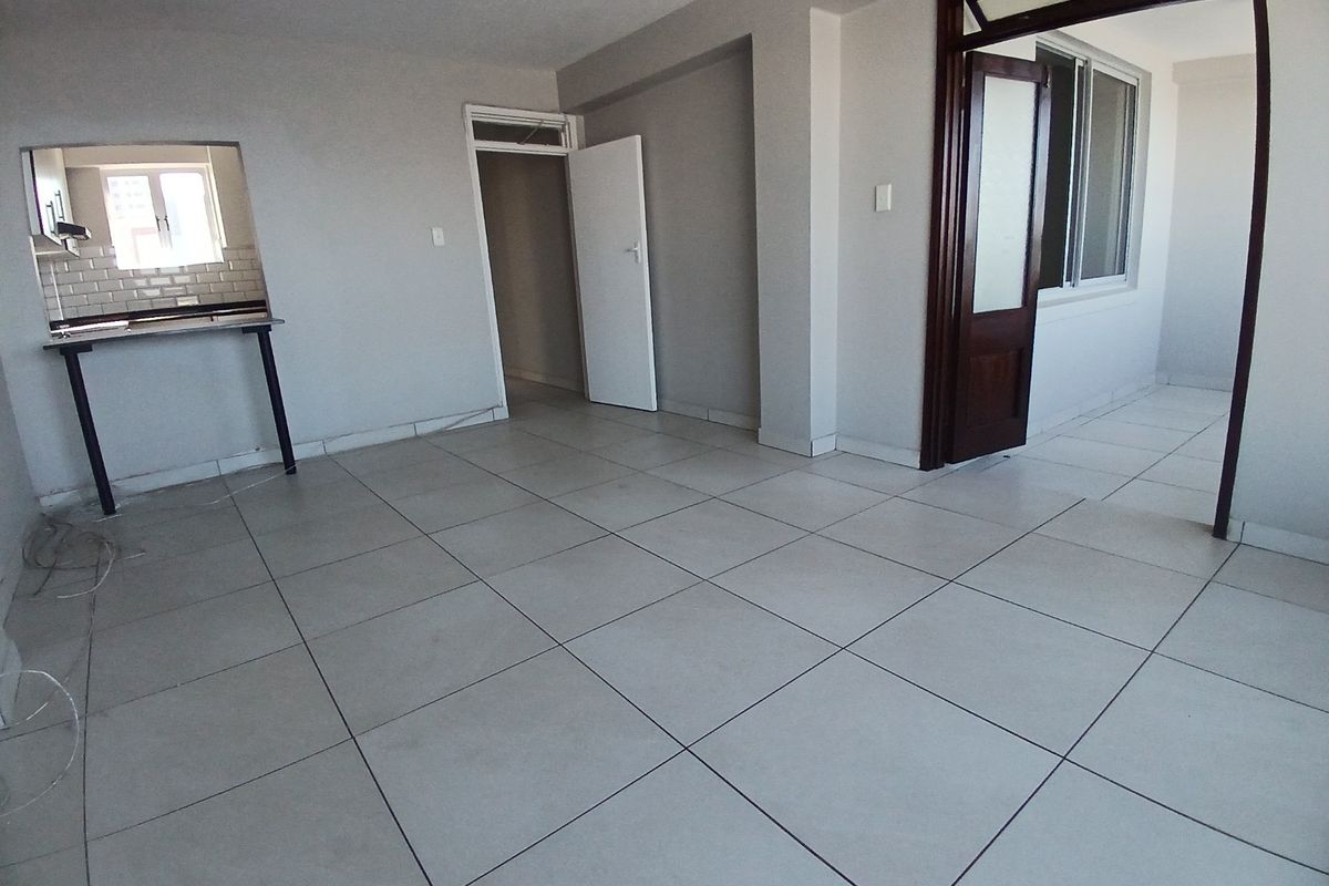 An empty open-plan living area featuring neutral tiled flooring, a kitchen pass-through window, and access to adjacent rooms.