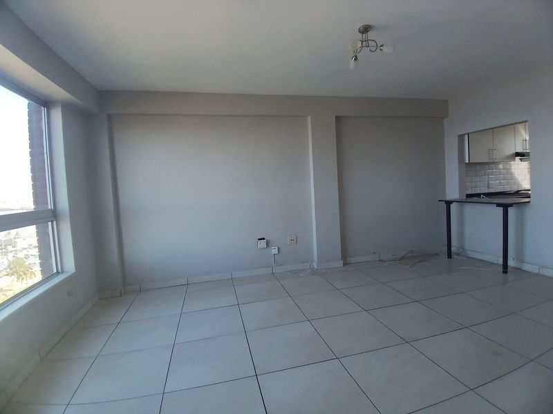 An empty open-plan living area featuring tiled flooring, a breakfast bar overlooking the kitchen, and large windows providing natural light.