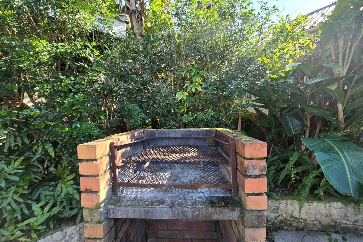 A built-in brick braai area situated in a lush, overgrown garden setting, featuring a weathered metal grill grate.