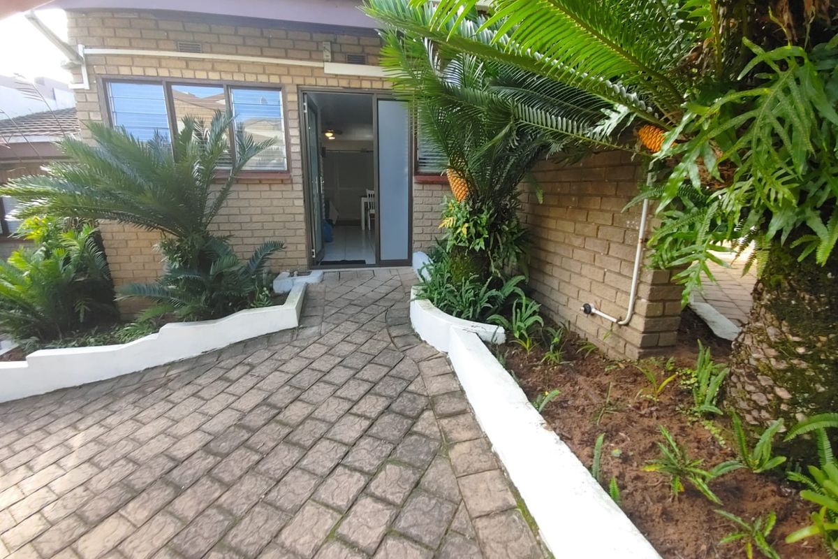 A rear exterior view of the townhouse featuring a paved patio area, brick facade, and established garden beds leading to a sliding glass entrance.