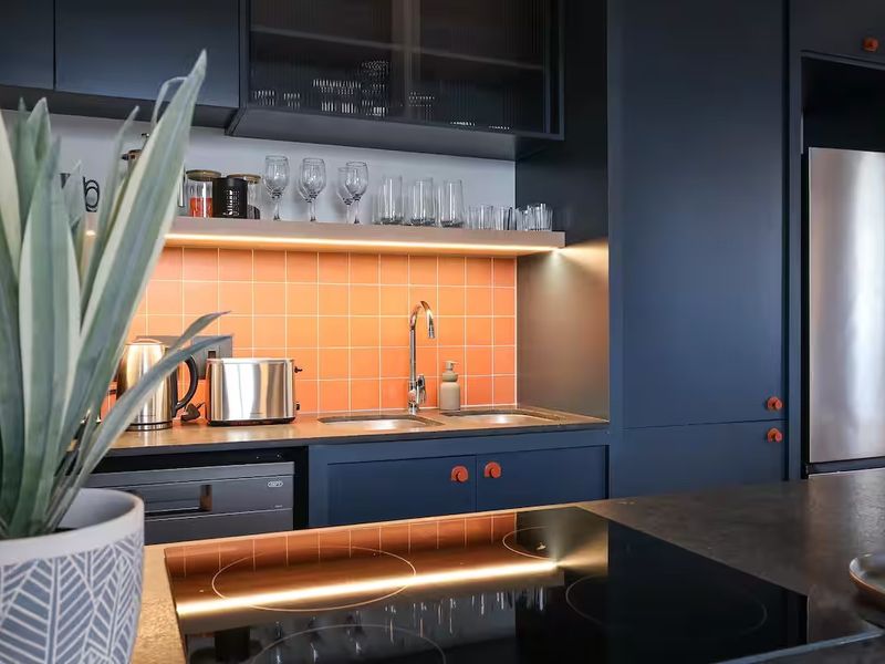 Modern kitchen featuring dark cabinetry, a vibrant orange tiled backsplash, integrated appliances, and a sleek induction cooktop with under-cabinet lighting.