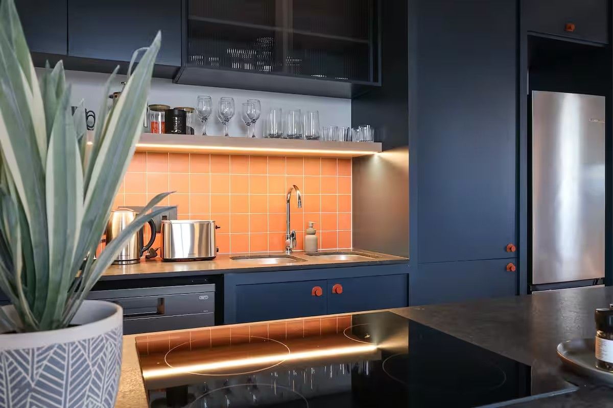 Modern kitchen featuring dark cabinetry, a vibrant orange tiled backsplash, integrated appliances, and a sleek induction cooktop with under-cabinet lighting.