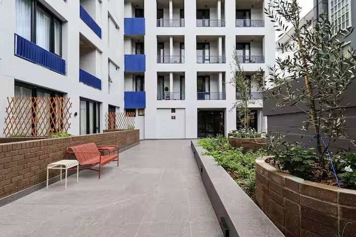 A well-maintained modern apartment complex courtyard featuring paved flooring, integrated brick planter boxes, and multi-level balconies with blue accents.