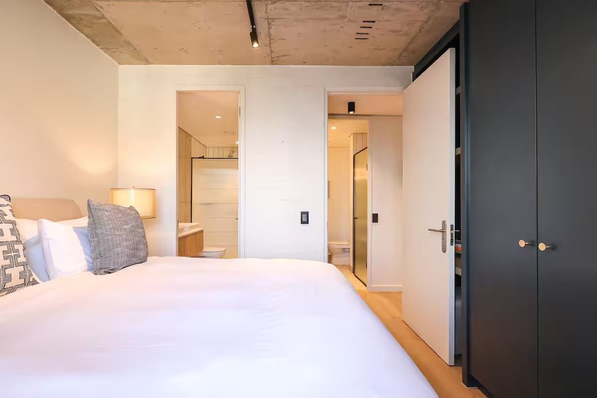 Modern bedroom featuring an exposed concrete ceiling, track lighting, and a sleek built-in wardrobe, with direct access to an en-suite bathroom.