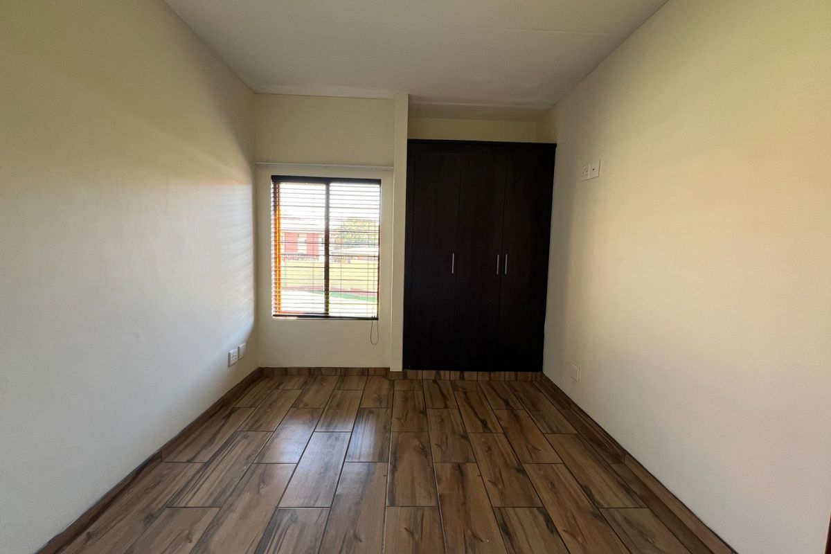 A clean, empty bedroom featuring wood-look tile flooring, a built-in dark wood wardrobe, and a window with fitted venetian blinds.