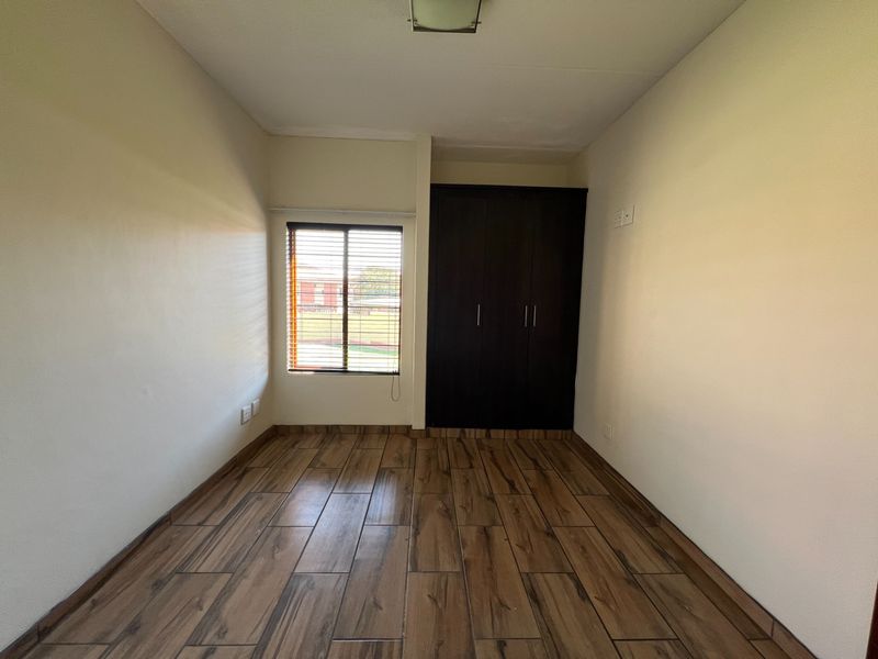 A clean, empty bedroom featuring wood-look tile flooring, a built-in dark wood wardrobe, and a window with fitted venetian blinds.