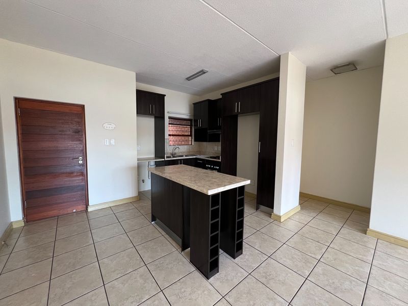 An open-plan kitchen featuring dark wood cabinetry, a central island with stone countertop, ceramic tiled flooring, and integrated cooking appliances.