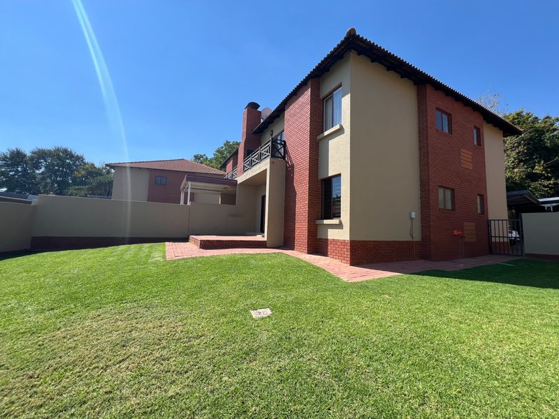 Rear view of the townhouse featuring a brick and plaster facade, a private balcony, a paved patio area, and a well-maintained lawn.