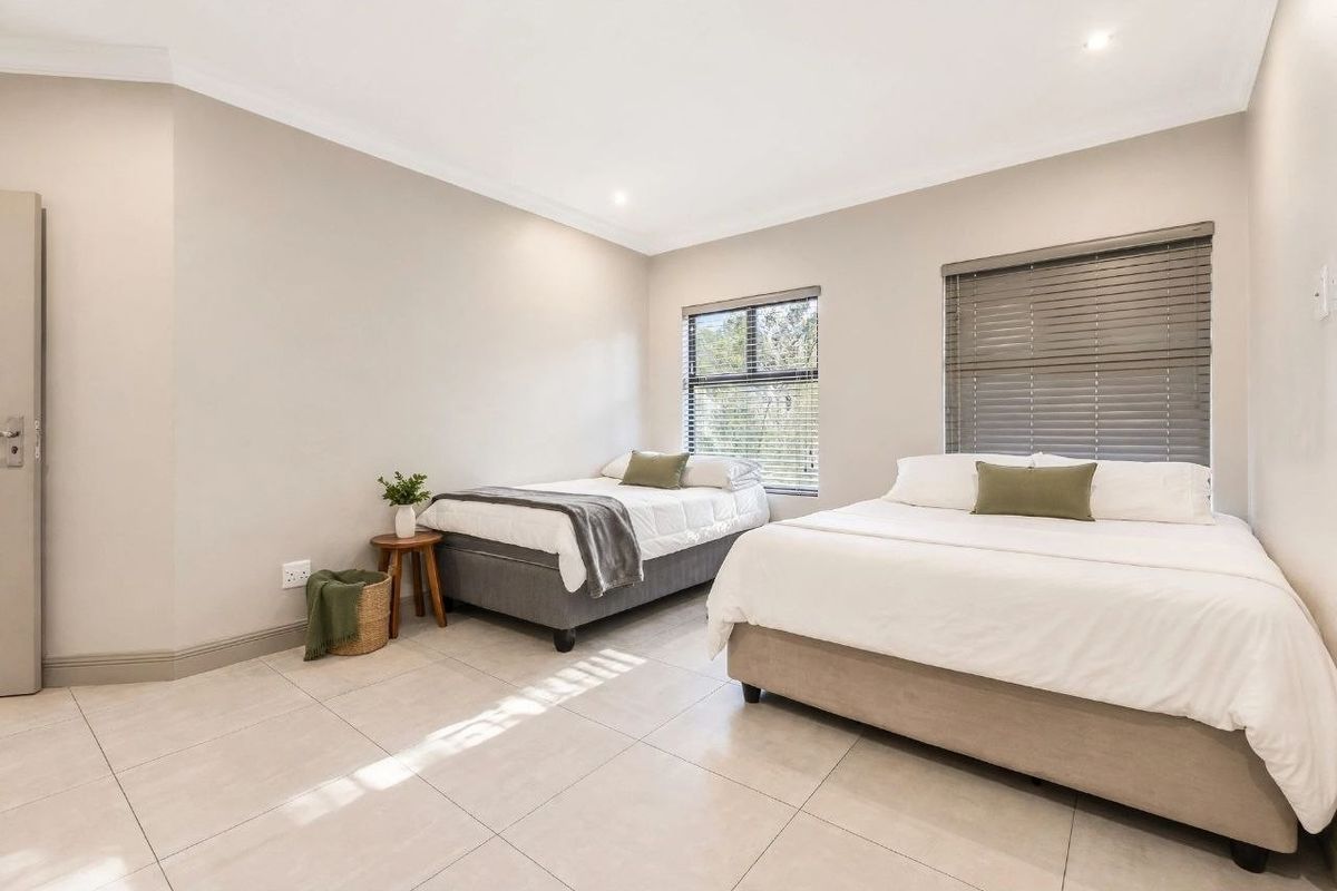 A spacious, well-lit bedroom featuring neutral tiled flooring, recessed downlighting, and large windows fitted with venetian blinds.