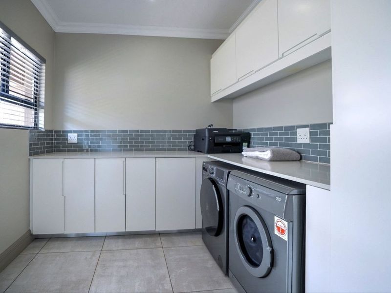 A modern, well-maintained laundry room featuring clean white cabinetry, stone countertops, a stylish grey subway tile backsplash, and large format floor tiles.