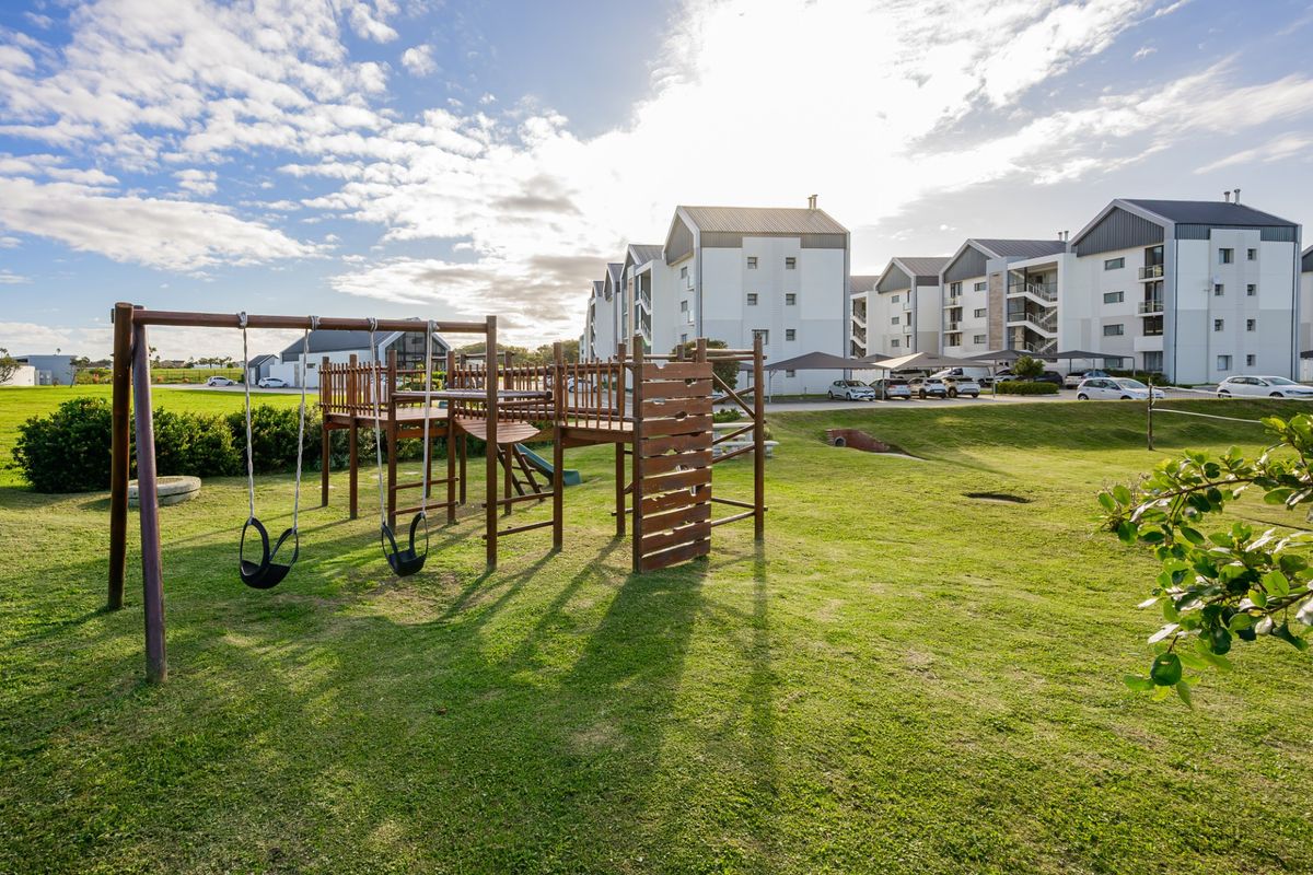 A well-maintained communal garden area featuring a wooden playground set, situated within a modern apartment complex with ample parking.