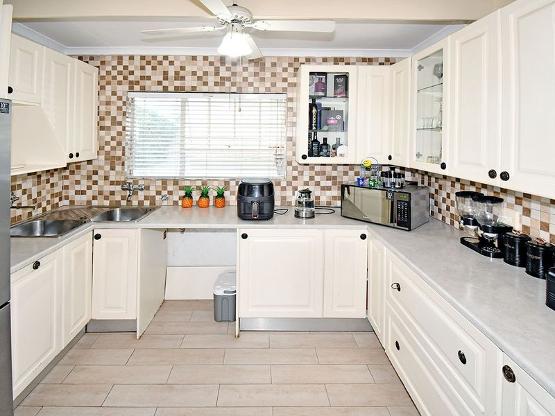 A functional kitchen featuring cream-colored cabinetry, tiled backsplash, laminate countertops, and a stainless steel double sink, with a ceiling fan for ventilation.