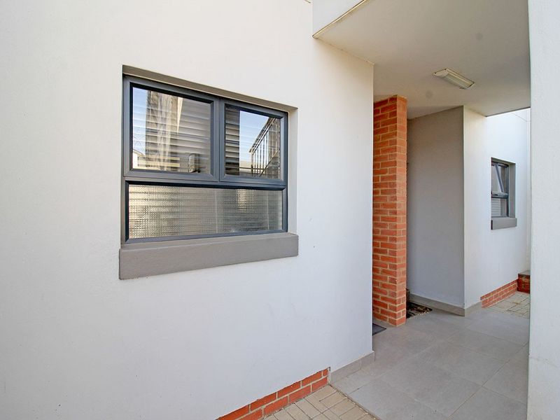 A clean and well-maintained apartment entrance featuring a covered walkway, brick accents, and modern aluminium window frames.