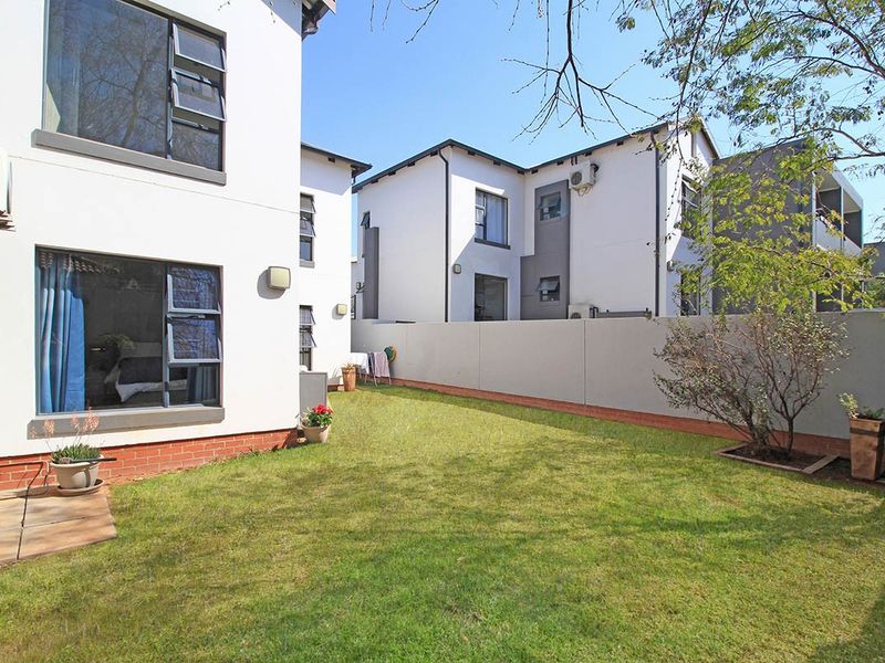 A private rear garden featuring a well-maintained lawn, paved patio area, and boundary wall, adjacent to the modern apartment building exterior.