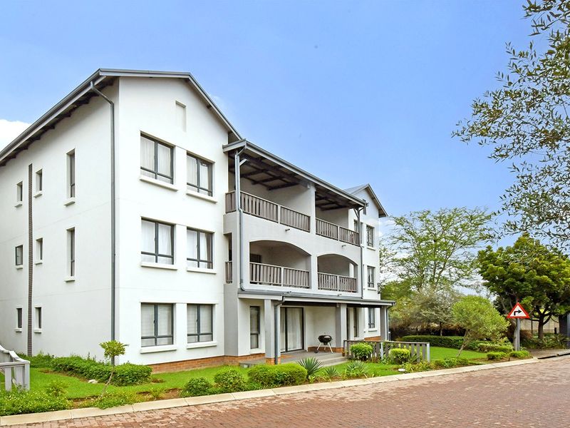 A multi-story apartment building featuring white facade, private balconies, and well-maintained surrounding garden areas with paved driveway access.