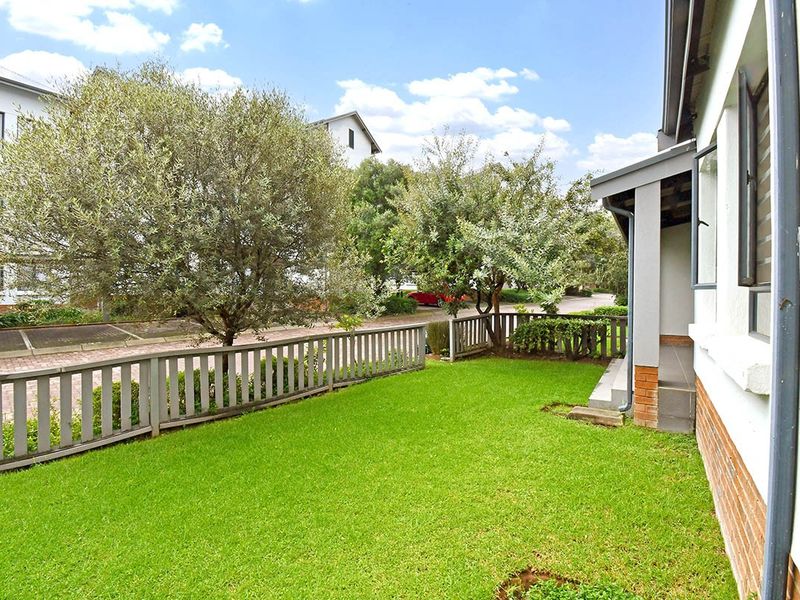 A well-maintained rear garden area featuring a lush green lawn, a wooden picket fence, and a covered patio attached to the apartment building.