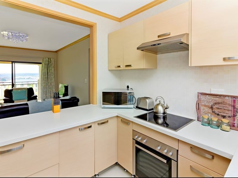 An open-plan kitchen featuring light-wood cabinetry, stone countertops, and integrated appliances, seamlessly flowing into a living area with balcony access and scenic views.