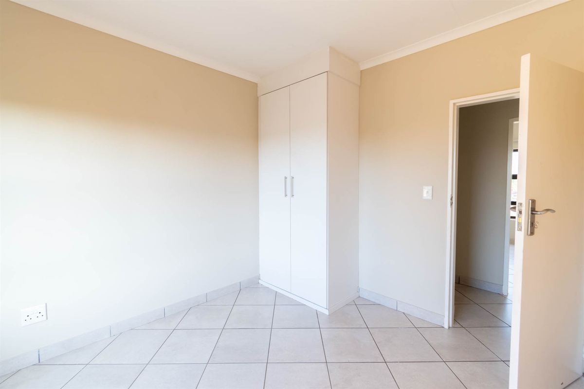 A clean, empty bedroom featuring neutral wall paint, tiled flooring, and a white built-in wardrobe with a doorway leading to an adjacent room.