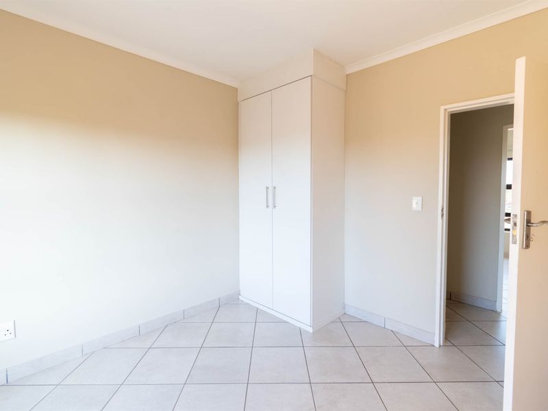 A clean, empty bedroom featuring neutral wall paint, tiled flooring, and a white built-in wardrobe with a doorway leading to an adjacent room.