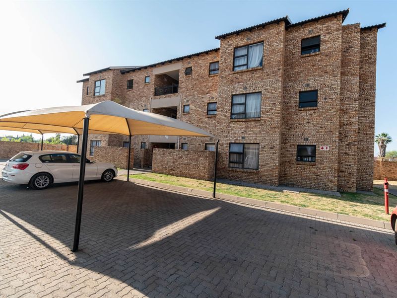 Exterior view of a multi-story face brick apartment complex featuring a paved parking area with shade cloth carports.