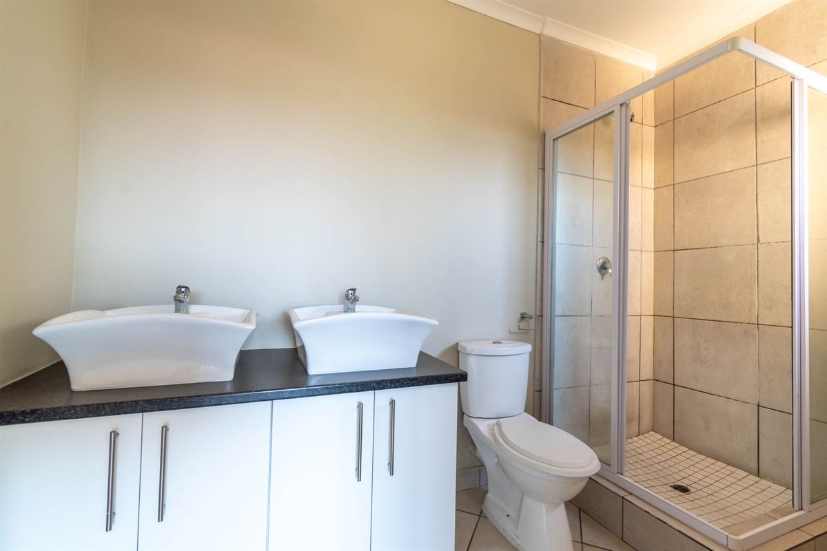 A clean, modern bathroom featuring a double vanity with white basins, a toilet, and a glass-enclosed shower with neutral wall tiling.