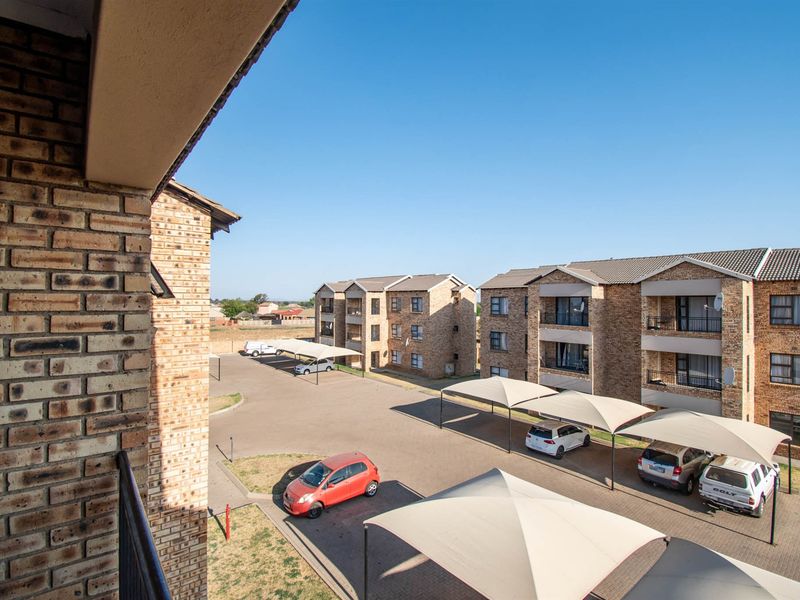 View from an elevated balcony overlooking the apartment complex grounds, featuring face brick buildings, paved parking bays, and shade cloth carports.