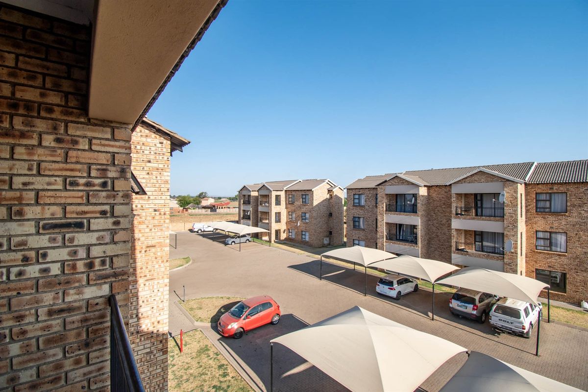 View from an elevated balcony overlooking the apartment complex grounds, featuring face brick buildings, paved parking bays, and shade cloth carports.