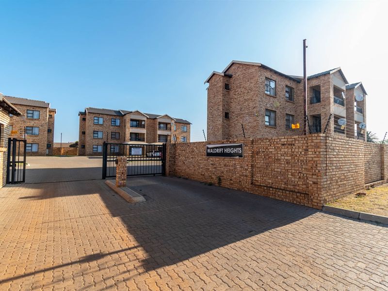 Exterior view of the Waldrift Heights apartment complex featuring face brick buildings, a paved driveway, and secure gated access with perimeter electric fencing.
