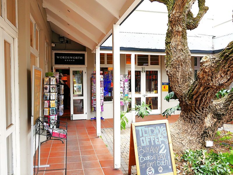 A covered outdoor walkway featuring terracotta tiling and glass-paned entrances, providing access to a retail storefront in a landscaped courtyard setting.