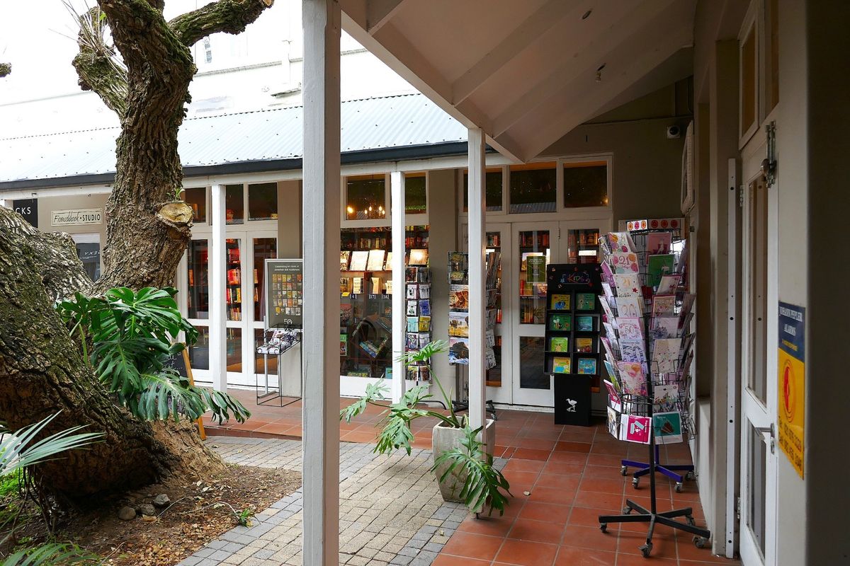 A covered outdoor walkway featuring terracotta tiling and timber support posts, providing access to retail storefronts with glass-paneled doors.