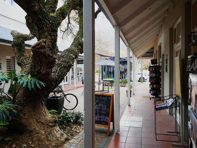 A covered outdoor walkway with tiled flooring and timber support posts, providing access to retail storefronts in a landscaped courtyard setting.