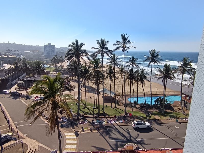 Elevated coastal view overlooking the beach, palm trees, swimming pool, and surrounding neighborhood infrastructure.