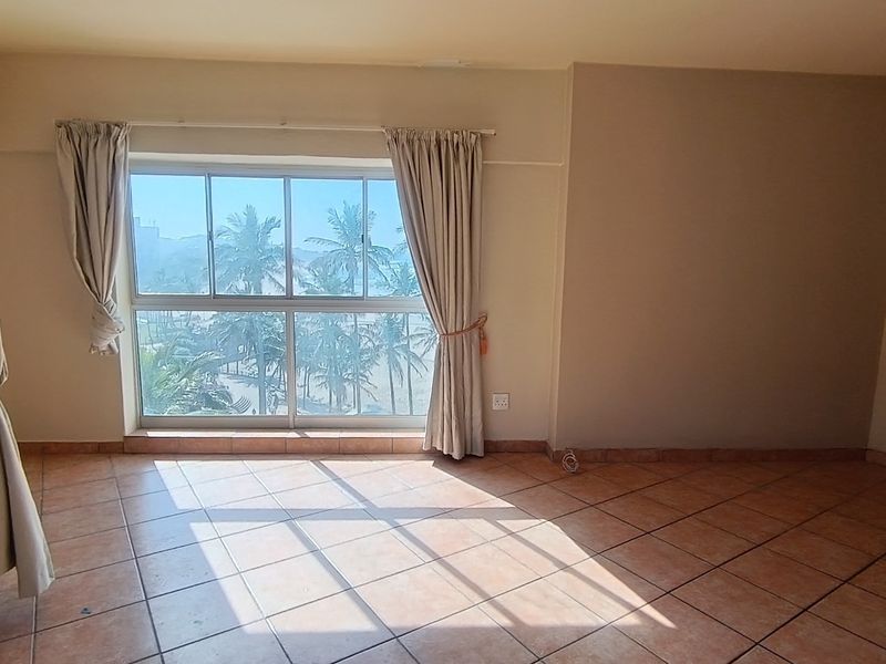 An empty, sunlit living area featuring tiled flooring and a large sliding window with views of palm trees.