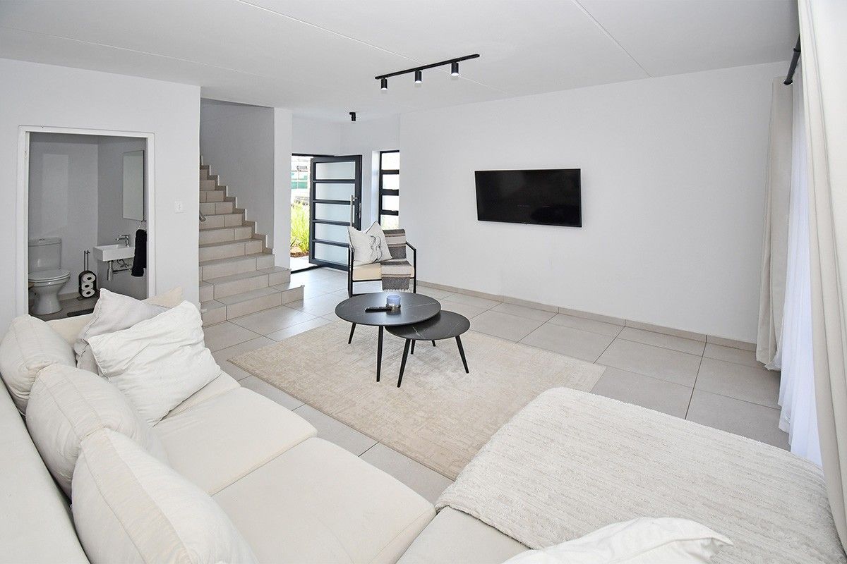 A bright, modern open-plan living area featuring neutral tiled flooring, track lighting, and a staircase leading to the upper level.