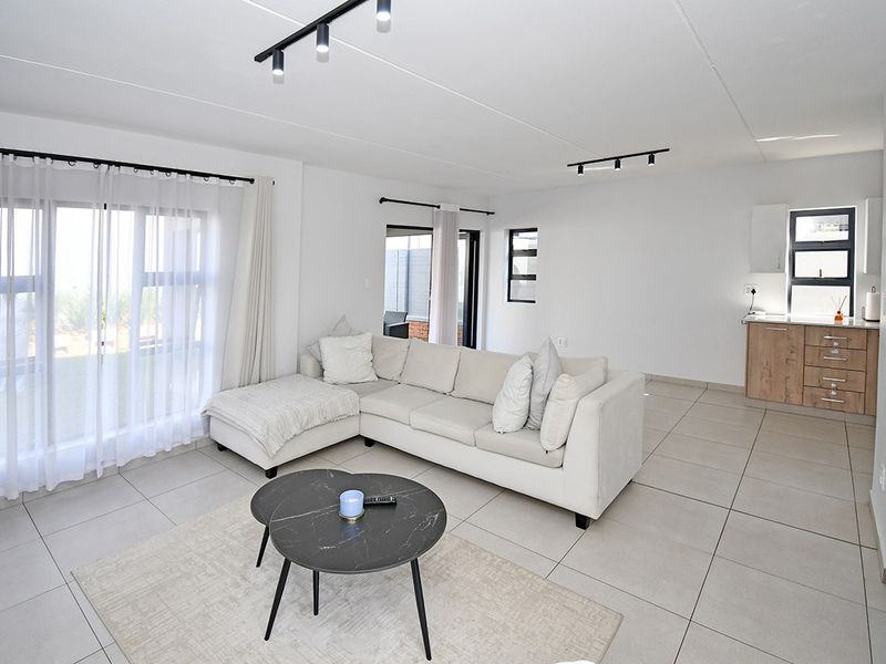 Modern open-plan living area featuring neutral tiled flooring, contemporary track lighting, and a compact kitchen with built-in cabinetry and stone countertops.