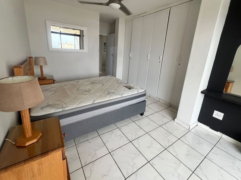 A bedroom featuring white tiled flooring, built-in cupboards, and a ceiling fan, offering a functional space with neutral finishes.