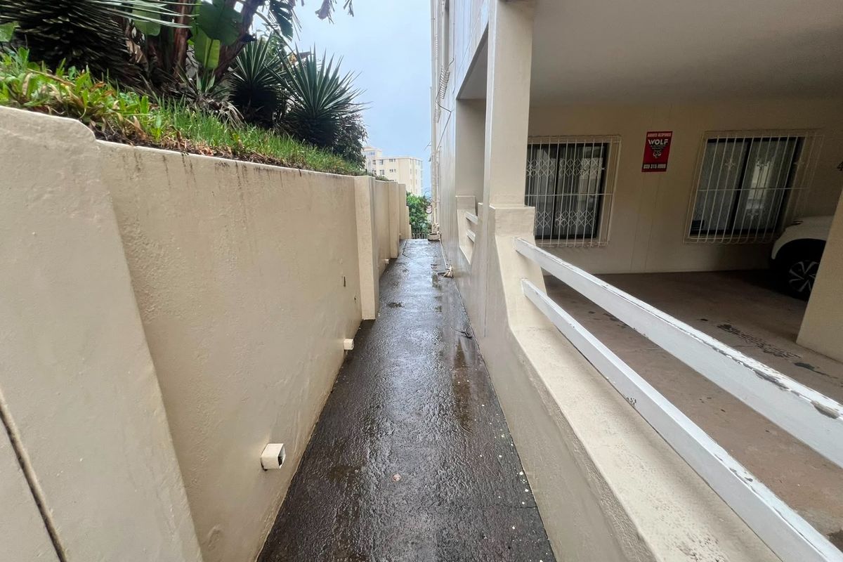 A narrow exterior concrete pathway running alongside a retaining wall and the building structure, featuring security bars on the windows.